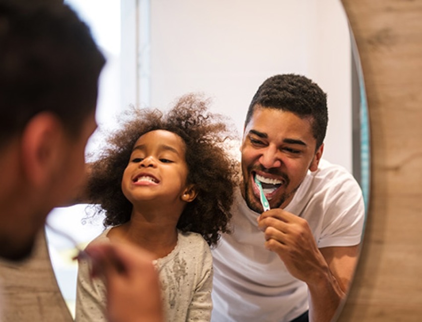 A man and a young girl are brushing their teeth together in front of a mirror, promoting dental hygiene and bonding.